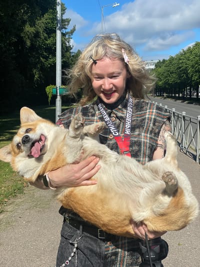 The media file shows a woman holding a corgi dog outdoors. The woman has blonde, wavy hair and is smiling widely, with her eyes closed. She is wearing a plaid shirt and has tattoos on her arm. A red strap with text is visible across her chest. The corgi dog is light brown and white, with its tongue hanging out and its mouth open in a happy expression. It is being held upside down by the woman, with its paws in the air.

The scene appears to be a park or outdoor recreational area, with trees and a paved path visible in the background. The sky is blue with some clouds, suggesting it is daytime and likely pleasant weather. A metal fence and a lamppost are also present. A small green planter with ivy is attached to the lamppost.

The visible text on the red strap appears to be in Cyrillic and says "СТИЛЬ БЕШЕНЫЙ", which translates to "MAD STYLE". Another word, "EXALIST", is visible in smaller font.

The overall mood of the image is joyful and playful, captured in the smiling faces of both the woman and the dog, and the dog's energetic pose. The setting suggests a casual outing on a sunny day.