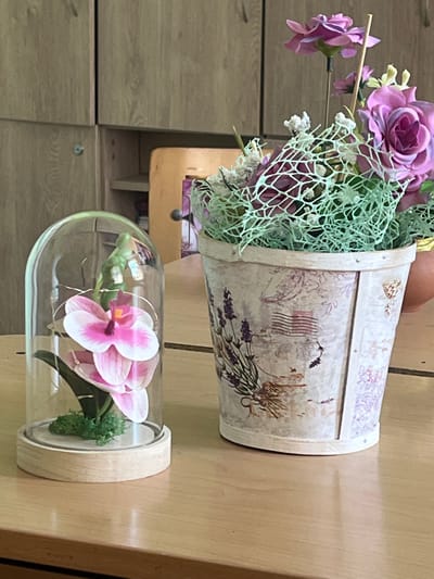 The image depicts a still life arrangement of two decorative floral displays on a wooden table. On the left, a single artificial pink and white orchid is encased in a glass dome with a wooden base, surrounded by decorative green moss and subtle fairy lights. To the right, a pot filled with artificial purple flowers, including roses and smaller blossoms, is adorned with a light green netting. The pot itself is decorated with a vintage-inspired pattern featuring lavender sprigs and postal stamps. In the background, a wooden cabinet with visible handles and shelves is partially visible, suggesting an indoor setting. The lighting suggests it is daytime. The scene is devoid of any individuals, actions, or interactions. The overall environment appears to be a domestic or decorative setting, possibly a home or a shop. The location of Cheshnegirovo, Bulgaria is not visually represented in the image.