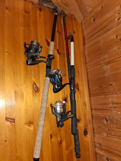 This image depicts a collection of three fishing rods with reels, vertically suspended against a wooden wall, likely in a storage area. The rods are arranged in a slight diagonal line. The leftmost rod is predominantly cork-handled with a black and silver reel. The middle rod appears to be a darker color with a blue and silver reel. The rightmost rod is black with a white section near the top and a black reel. The wooden wall exhibits a visible grain pattern and natural knots. The lighting suggests an indoor environment. The scene is static, with no people or action present. The location is identified as Åsgårdstrand, Norway.