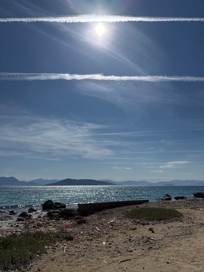 Bright sunlight illuminates a coastal scene in Aegina, Greece. The sky is clear blue with a prominent sunburst and lens flare in the upper right quadrant, accompanied by two distinct, parallel white contrails traversing horizontally. Wispy cirrus clouds are also visible throughout the upper sky. The Aegean Sea occupies the midground, its surface characterized by extensive sparkling reflections from choppy water. A distant, dark mountainous landmass is silhouetted against the horizon. The foreground presents a transition from a rocky shoreline on the left to a sandy and gravelly beach on the right. A low, eroded concrete or stone structure, possibly a dilapidated pier or breakwater, extends from the beach into the water on the right. Sparse green vegetation patches are present on the sandy ground, where a small red object, appearing to be plastic debris, lies on the gravel near the center. Additional scattered small debris is visible on the sand and rocks. No human activity or individuals are observed within the frame.