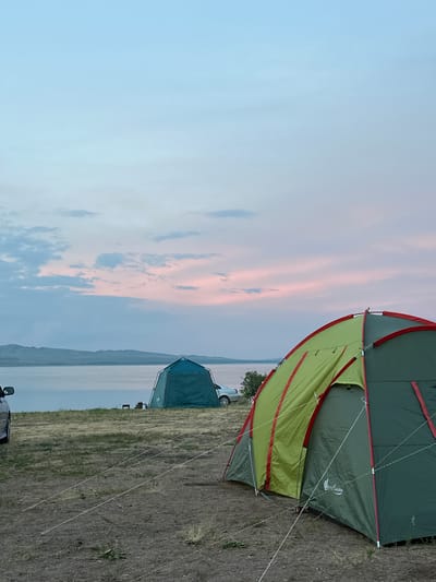The image depicts a serene campsite at what appears to be either dawn or dusk, given the soft, pastel hues of the sky and the gentle illumination. Two tents are set up on dry, grassy terrain. The foreground is dominated by a large, green and lime-colored tent with red trim, partially open to reveal an inner sleeping compartment. This tent is secured by numerous guy lines, suggesting it is a robust structure designed to withstand elements. In the background, a smaller, dark teal tent is visible, positioned closer to a body of water.
The scene overlooks a vast expanse of water, possibly a lake or a sea, with a low-lying landmass or distant mountains forming the horizon line. The sky is painted with wispy clouds in shades of pale pink, soft blue, and muted gray, indicating a calm atmospheric condition. A silver car is parked near the larger tent, and another vehicle, possibly a black SUV, is partially visible in the lower left corner. The ground around the tents is covered in dry grass and scattered small plants.
The overall mood is peaceful and tranquil, evoking a sense of calm and connection with nature. The time of day suggests a quiet transition between day and night, a popular time for campers to relax and enjoy the scenery. No people are explicitly visible in the image. There is faint text on the larger tent, appearing to be a logo or brand name, possibly including the word "Nordisk" or a similar stylized script, though it's difficult to read clearly due to resolution and angle. The location context of Kolodeznyy, Russia, places this scene within a natural, possibly rural or semi-wild environment.