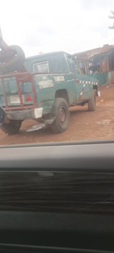 The image shows a green pickup truck parked on a dirt road in Ozalla, Nigeria. The truck has a custom rear rack with a tire mounted on it. A red triangle is visible on the back. The truck's body has faded paint and what appears to be some reflective striping along the side. Some type of building is behind the truck. The image was taken from inside a vehicle, as a portion of the dashboard is visible in the foreground. The overall environment appears to be rural or semi-rural.