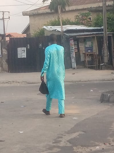 A man in a light blue traditional outfit walks away from the camera, holding a black bag. The background includes a building with a metal gate, a small shop-like structure, utility poles, and various buildings. The scene is set outdoors during daylight hours. The road the man walks on shows signs of wear and has scattered debris.