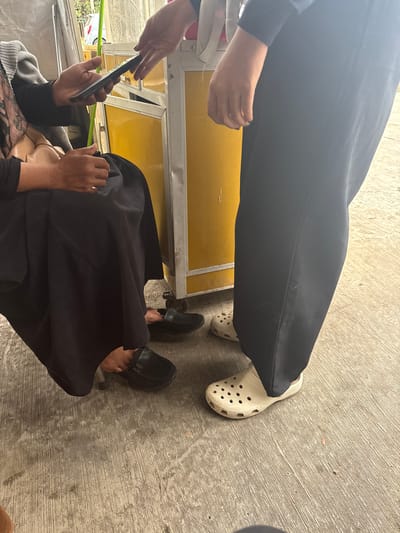 In Meunasahmanyet, Indonesia, two individuals are engaged in an exchange, with one person on the left seated and holding a mobile phone, while the other person on the right stands and reaches out. They are positioned near a yellow food cart with a metal frame. The ground is concrete, and the setting appears to be outdoors. The seated individual wears a black abaya and black slip-on shoes, while the standing individual wears a long black garment and light-colored Crocs.