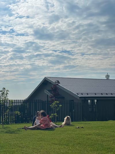 In Simagino, Russia, on a clear day with scattered clouds, three people are seated on a black blanket on a well-maintained green lawn. Two individuals are in conversation, while the third person appears to be looking away. A light-colored, fluffy dog is situated to the right of the group, facing the camera. Behind them, a dark wooden fence encloses the property, and a modern house with a grey metal roof stands in the background. Trees and shrubbery are visible around the property.