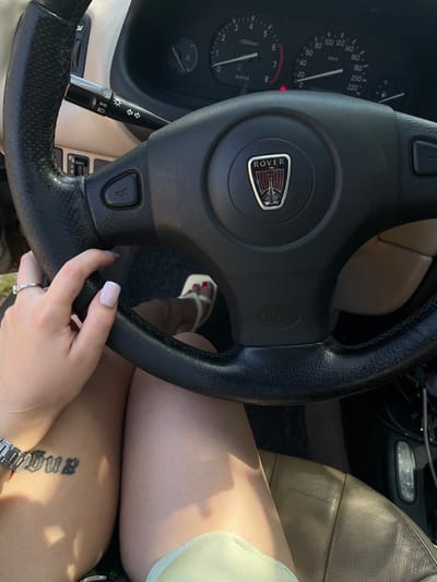 The image shows a close-up view of a person's hands and legs inside a car. The steering wheel, dashboard with a speedometer and tachometer, and part of the car seat are visible. The person is wearing a ring on their left hand and has a tattoo on their left thigh. The car appears to be a Rover, as indicated by the logo on the steering wheel. The lighting suggests it is daytime. There is no indication of the location in the city of Пліскі сельскі Савет, Belarus.