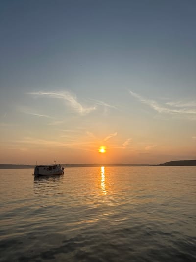 The image depicts a serene sunset over a body of water, likely a lake or a wide river, with a small white boat anchored in the foreground to the left. The sky transitions from a pale blue at the top to a vibrant orange and yellow near the horizon, where the sun is setting. Wispy clouds are scattered across the sky, catching the warm hues of the sunset. The water reflects the colors of the sky and the sun, creating a shimmering, golden path. A faint, dark outline of a distant shoreline with trees is visible across the water. The overall atmosphere is tranquil and peaceful. The image is set in the city of Sylva, Russia.