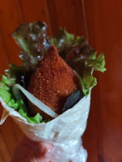 A hand holds a wrap filled with food. The wrap is made of a thin, white flatbread, twisted to hold the contents. Inside, a fried, conical-shaped food item, likely a croquette or empanada, is prominently displayed. It has a golden-brown, crispy exterior. Surrounding the fried item are fresh green lettuce leaves, some ruffled and some smoother, and a few dark green herb leaves, possibly basil. There are also thin, white slices, which could be onion or another vegetable. The background is a warm, reddish-brown wooden surface, suggesting an indoor setting. The lighting appears to be natural or soft indoor light, casting gentle shadows and highlighting the textures of the food. There is no visible text. The focus is entirely on the food, suggesting it is being presented or eaten. The overall impression is of a homemade or street food item, ready to be consumed.