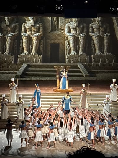 A staged scene of what appears to be an ancient Egyptian opera, displayed indoors. A massive backdrop resembling temple architecture with giant seated figures flanks the left of the frame. Center stage are a pair of figures elevated on a platform, costumed in blue and white. Numerous other figures, similarly costumed, kneel and raise their arms toward the elevated figures. Other figures stand on either side of the kneeling ones, holding what appear to be staffs. The scene suggests a performance or ceremony.

The image does not include information about the location in the city of San Borja, Peru.