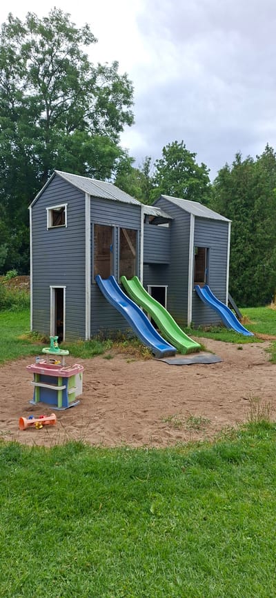 The image depicts an outdoor playground featuring a dark gray, two-story play structure with corrugated metal roofs. The structure has multiple openings, including doorways and windows, and is equipped with three slides: two blue and one green, all positioned on the front of the playhouses. The ground in front of the structure is covered with sand, and some grass is visible in the foreground and background. Scattered on the sand are a pink and blue toy kitchen set and a small orange toy. The background is dominated by lush green trees and an overcast sky. The location is identified as Kata, Estonia. No people are visible in the image.