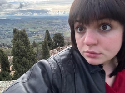 An image displays a young female, positioned on the right half of the frame, looking directly at the camera with wide-open eyes and slightly parted lips. She has short, dark hair with bangs and is wearing a black leather jacket over a red garment. Her left shoulder and upper arm are visible. In the background, from an elevated viewpoint characteristic of Cortona, Italy, a panoramic view of the Val di Chiana valley unfolds. This landscape features extensive green agricultural fields, scattered residential structures, and numerous dark green cypress trees. Red-tiled rooftops of buildings are visible immediately below the viewpoint. A distant body of water, likely Lake Trasimene, is discernible on the horizon under a cloudy sky. A textured stone wall or ledge is partially visible in the lower left corner.