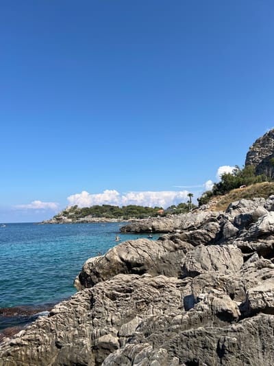 A coastal scene in Palermo, Italy, features a rocky cliff face on the right side of the frame, jutting into a body of clear, blue water. The water has gentle ripples and some people can be seen swimming and kayaking. Beyond the water, a lush green coastline is visible, dotted with buildings and trees, suggesting a populated area. The sky above is a vibrant, cloudless blue, indicating a sunny day. The lighting suggests it is midday. The overall mood is one of leisure and natural beauty.