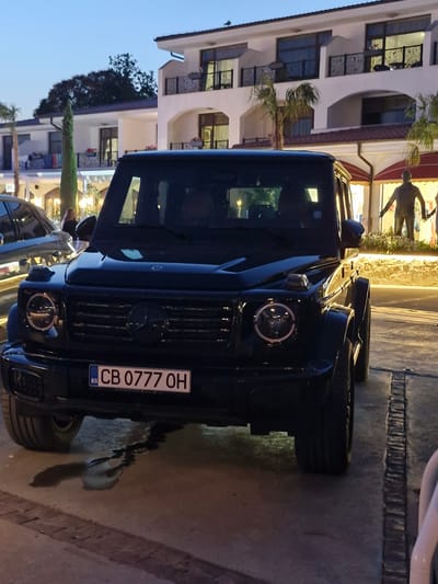 The image shows a black Mercedes-Benz G-Wagon parked on a paved surface in front of a multi-story white building with arched windows and balconies. The building appears to be a hotel or resort, and there are palm trees and other greenery visible. A statue of a man is visible in front of the building, to the right of the car. The license plate of the car reads "CB 0777 OH". There is a puddle of liquid on the ground beneath the front of the car. The sky is a dusky blue, suggesting it is either early morning or late evening. The location is the resort town of Sveti Vlas, Bulgaria.