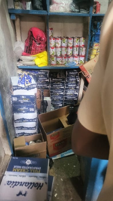 The scene depicts an interior retail or storage area in Fagge D1, Nigeria, characterized by stacked packaged goods. A blue wooden shelving unit occupies the left and central portions of the frame. The top shelf contains miscellaneous items including clear plastic bags and small packages. The middle shelf features a red plastic bag, a yellow plastic bag, and a white plastic strainer/basket on the left, while the right section holds approximately 30-40 cans, predominantly with red, white, and green labels, stacked in multiple rows (likely Hollandia evaporated milk). The bottom shelf and the floor are extensively stocked with numerous dark blue cardboard boxes, clearly labeled "Hollandia Full Cream Evaporated Milk," arranged in dense stacks. Several open brown cardboard boxes, some labeled "RIVO Original," are also visible on the floor, some containing additional cans. A single individual is partially visible on the far right, with only their right shoulder and arm, clad in a light-colored shirt, entering the frame. The environment features light-colored walls and a concrete floor, with ambient indoor lighting.