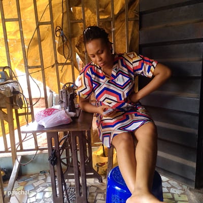 The image shows a woman seated in what appears to be a tailoring workspace in Nsugpe, Nigeria. She is fair-skinned, with dark hair adorned with small, white decorative beads. She's wearing a short-sleeved dress with a geometric pattern of red, blue, white, yellow, and black. She is holding a mobile phone and looking down at it. She has one hand on her hip.
She is seated on a bright blue plastic stool. To her left is a vintage-style sewing machine sitting on a dark metal table with a plastic bag of red and white fabric next to it. A yellow fabric hangs behind a metal-barred window. To the right is a dark, corrugated metal wall. The floor appears to be tiled with irregularly shaped pieces. The name "paschal" is visible in the lower-left corner of the image.