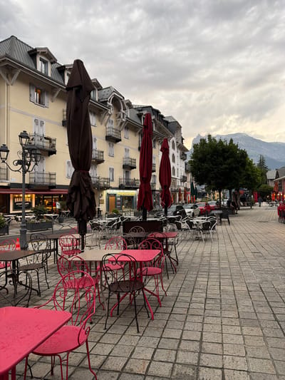 The image depicts a street scene in Saint-Gervais-les-Bains, France, with an outdoor seating area in the foreground. Several tables and chairs, some painted bright pink, are arranged on a cobblestone pavement. Large closed umbrellas, one dark brown and others red, stand prominently. Behind the seating area, a building with cream-colored walls, multiple balconies, and grey roofs lines the street. Shops with signage, including "BRASERIE MONT BLANC" and "LA FERME DU MONT BLANC," are visible on the ground floor of the building. Further down the street, trees and more buildings are present, with a mountain range visible in the background under a cloudy sky. The pavement appears wet, suggesting recent rain. No people are actively engaged in actions or interactions within the frame, though figures can be seen seated at tables in the distance.