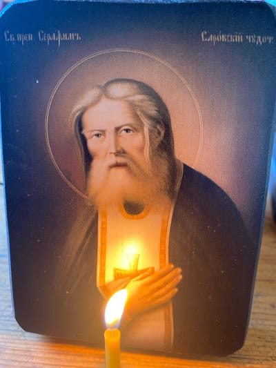 A religious icon depicting an elderly man with a long beard and flowing hair, likely Saint Seraphim of Sarov, is illuminated by a lit candle. The icon has Cyrillic text above and to the right of the figure, which reads "Св. преп. Серафимъ" and "Саровскій чудот." respectively. The icon is placed on a wooden surface, possibly a table. The background of the icon is dark blue, with a halo around the saint's head. The candle is yellow and has a bright flame. The image appears to have been taken in Zelenodol'sk, Russia, as indicated by the context.