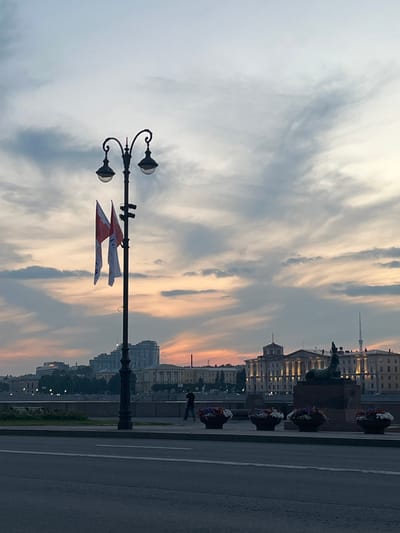 This image depicts a waterfront scene in the city of Ligolambi, Russia, during twilight. A prominent lamppost with two lights stands in the foreground, adorned with two flags. In the background, across a body of water, lies a cityscape featuring various buildings, including one with classical architecture illuminated by lights. A statue, possibly of an animal, is visible on a pedestal in front of the illuminated building. Along the waterfront promenade, planters filled with colorful flowers are arranged in a line. A lone figure walks along the promenade. The sky above is filled with dramatic clouds, tinged with the warm hues of sunset. The overall atmosphere is serene and picturesque.