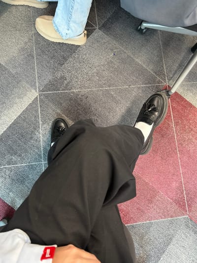 The image presents a top-down view of the lower bodies of two individuals seated indoors on a patterned carpet. The primary subject, in the foreground, wears black trousers, white socks, and black chunky-soled loafer-style shoes adorned with tassels. Their legs are crossed, and a white shirt cuff with a red rectangular tag is visible on the lower left. A second individual is partially visible in the upper left, wearing light blue jeans and tan slip-on shoes. The floor is covered by a carpet featuring a geometric design with sections of dark gray, lighter gray, and a prominent red triangular area on the right. A silver metal leg, consistent with a chair or table, is visible on the right side, positioned on the red carpet section. The specific location within Garston, United Kingdom, cannot be determined from the provided visual information.