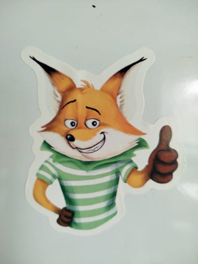 A sticker of a cartoon fox wearing a green and white striped shirt and a green collar is displayed on a white surface. The fox has orange fur, large ears with black tips, and wide, expressive eyes. It is smiling broadly and giving a thumbs-up with its right hand. The background appears to be a wall or a board, possibly in a home or office setting. There is no discernible location in the city of Kryvaselski Sel'ski Savet, Belarus.