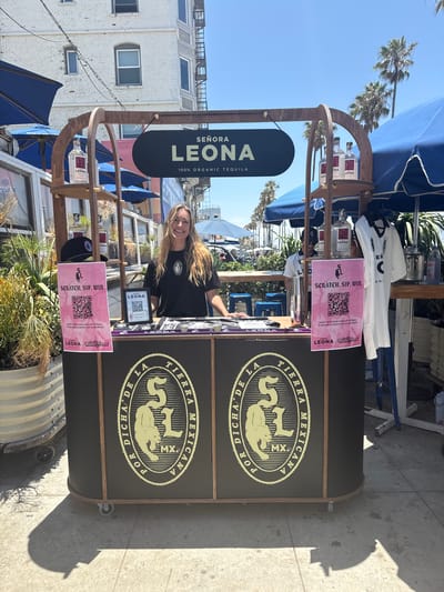 This is a medium shot photograph of a woman standing behind a wooden booth in Santa Monica, California. The booth is dark brown with a curved wooden frame. A black oval sign hangs from the frame and reads "SEÑORA LEONA 100% ORGANIC TEQUILA" in white lettering. The booth has two shelves on the left side, displaying three bottles of "LEONA" tequila, and a black baseball cap. A pink sign with a QR code and text "SCRATCH. SIP. WIN." is attached to the left shelf.

The woman is wearing a black t-shirt with a white logo. She has long blonde hair and is smiling at the camera. She appears to be interacting with customers.

The booth's front panel has a large circular logo with the words "POR DICHA DE LA TIERRA MEXICANA" and "SL MX" in a stylized font. To the right of the woman, a white t-shirt with a black logo is displayed. Various items, possibly merchandise, are arranged on the counter.

The background shows a bright, sunny day with a clear blue sky. Palm trees are visible in the distance, suggesting a coastal location. A multi-story white building with windows and a fire escape stands behind the booth. Blue umbrellas are set up to the left and right of the booth, providing shade. The ground is paved with concrete. A large planter with dry vegetation is positioned to the far left.