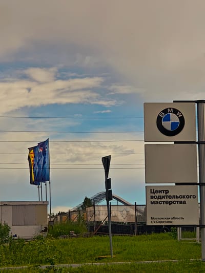 The image depicts a BMW dealership or service center in Il'inskoe, Russia, under a cloudy sky. A prominent BMW logo is displayed on a white sign, with the text "Центр водительского мастерства" (Center for Driving Skills) and "Московская область г/к Сорочаны" (Moscow Oblast, Sorochany complex) below it. In the background, several flags are visible, including a yellow and red flag and two blue flags. There are also utility wires crossing the frame, and a fence with construction materials and greenery behind it. A white modular building is visible on the left side of the image. The overall scene suggests a business establishment in a somewhat industrial or semi-rural setting.