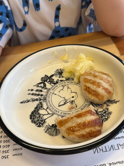A close-up shot shows a white ceramic plate with a black rim containing two grilled meat patties and a portion of mashed potatoes. The plate features a decorative black illustration of floral elements and a rabbit. To the bottom left of the plate, a menu with Russian text and prices is partially visible. The background includes a wooden table and a person wearing a white t-shirt with a blue and black animal print. The overall scene suggests a casual dining setting, likely a cafe or restaurant. The specific location in Tomsk, Russia, cannot be determined from the visual information provided.