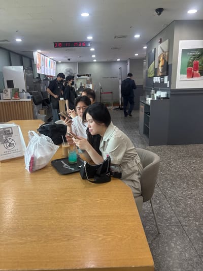 In a brightly lit interior that appears to be a cafe or similar establishment in Jeju City, South Korea, three individuals are seated at a wooden table. In the foreground, a woman with dark hair wearing a light-colored outfit is looking down at her phone, which she holds in her right hand. Beside her, a man also gazes at his phone. To their left, a third person, partially obscured, is also engaged with a phone. On the table are drinks, including a blue beverage and a coffee, a black tray, a white plastic bag, and a black purse. In the background, cafe staff in uniform are visible behind a counter, and a digital clock on the ceiling displays "3 18 11 17 3 16". Posters advertising "SEASON JUICE" are displayed on the wall. The overall environment suggests a casual setting where patrons are using their mobile devices.