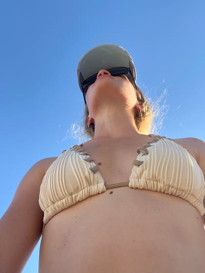 The image shows a low-angle, close-up shot of a person wearing a light-colored bikini top and a baseball cap, looking upwards against a clear blue sky. The person's face is partially obscured by the cap and sunglasses. The background is entirely sky, indicating an outdoor setting with no visible city landmarks or urban elements from Miljevci, Montenegro.