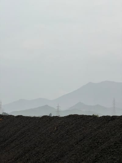 The image depicts a large pile of dark, granular material, possibly coal or industrial waste, in the foreground. Beyond this pile, a series of hazy, layered mountains are visible under a muted, overcast sky. Several electricity pylons are scattered across the landscape, with one prominent pylon to the right of the frame and another to the left. The overall atmosphere is somber and industrial, with a lack of discernible human presence or activity. The scene is likely situated in or near Saphale, India, given the mountainous terrain and the presence of industrial infrastructure, though no specific identifying landmarks are present to confirm the exact location.
