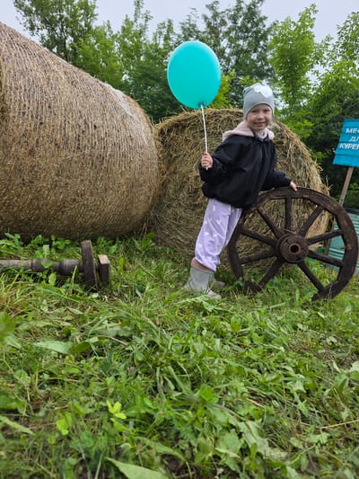 A young girl stands outside next to two large hay bales and a wooden wagon wheel. The girl is wearing a gray hat with a sparkly design, a black jacket with a pink fuzzy lining, and light purple pants with heart patterns. She is smiling and holding a bright turquoise balloon on a string. To her left is a large, round hay bale, and behind her is a second, smaller hay bale. A dark wooden wagon wheel is leaning against the smaller hay bale. The setting appears to be outdoors, likely a farm or rural area, with lush green grass and trees in the background. The lighting suggests it might be daytime, possibly overcast. A blue sign with white text is visible in the upper left corner. The overall mood appears cheerful and playful. The visible text on the sign reads "МЕСТ ДЛЯ КУРЕНИЕ" which translates to "SMOKING AREA".
