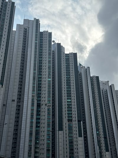 This media file shows a close-up, low-angle shot of a modern, tiered skyscraper with a repeating facade of windows and architectural elements. The building's design features sharp, angular lines and a stepped structure, creating a sense of immense scale and height. The exterior is primarily composed of grey and white panels, with horizontal bands of windows that are reflective.

A green helicopter is visible on a ledge or platform about midway up the building, suggesting maintenance or specialized access. The sky above is heavily overcast with dramatic, bright clouds that partially obscure the sun, indicating a cloudy or potentially stormy day. The overall mood is somewhat dramatic and imposing due to the scale of the building and the dramatic lighting conditions.

The image is taken from below, looking up at the structure, which emphasizes its verticality. The building's complex geometry and repetitive patterns of windows create a visually striking, almost abstract pattern. The location is identified as Hong Kong, China, a city known for its dense urban landscape and numerous skyscrapers. No visible text is present in the image. The time of day appears to be daytime, given the illumination from the sky.