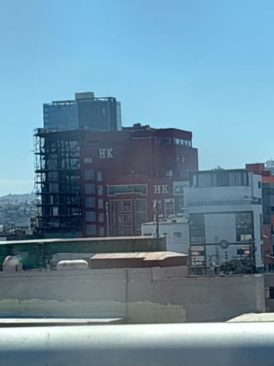 The image depicts a view of the Hong Kong building in Tijuana, Mexico. The building, characterized by its red facade with "HK" lettering, is captured from a slightly elevated position, likely from within a building or structure. The surrounding cityscape features other buildings of varying heights and architectural styles. The sky is clear and bright.