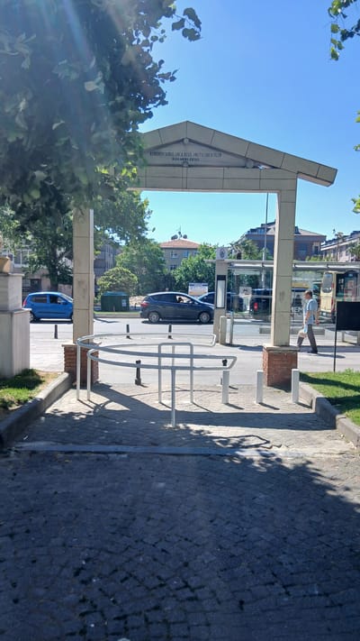 The image shows an outdoor scene in the Bahçelievler district of Istanbul, Turkey. A large, arched concrete structure with text inscribed on it is visible in the background, partially obscured by trees. The text on the structure reads "KOMANDO VURULUNCA DEGIL UNUTULUNCA OLUR RUHLARINA FATIHA." In the foreground, a white metal barrier structure, possibly a bike rack or turnstile, stands on a cobblestone path. Two cars, a blue hatchback and a dark gray hatchback, are driving on a street behind the barrier. A bus stop shelter with advertisements is also present. A man wearing a light blue shirt and dark pants is walking on the sidewalk, carrying a light-colored bag. The environment is sunny with clear blue skies and trees with green leaves. The ground is paved with cobblestones and some grass is visible. The overall scene depicts a public urban space in Bahçelievler.