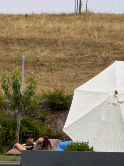 In Junglinster, Luxembourg, a person is captured relaxing outdoors. The scene is set in a grassy area, possibly a garden or park, with a large white umbrella providing shade. The individual, wearing a dark tank top with yellow accents and light-colored shorts, is seated on a low structure, possibly a chair or bench, with their legs crossed. One foot, clad in a red slipper, is visible.
The background consists of dry, golden-brown grass, suggesting a warm, possibly summery, day. Lush green foliage and bushes are interspersed within the landscape, and a bright blue object, perhaps a planter or a piece of garden furniture, is partially visible. The perspective of the image, looking up from a lower angle, hints at a possibly casual and relaxed atmosphere. The person appears to be looking towards the camera, their expression neutral or slightly intrigued. The overall lighting suggests daytime, with the sun likely high in the sky, casting soft shadows. There is no visible text in the image.