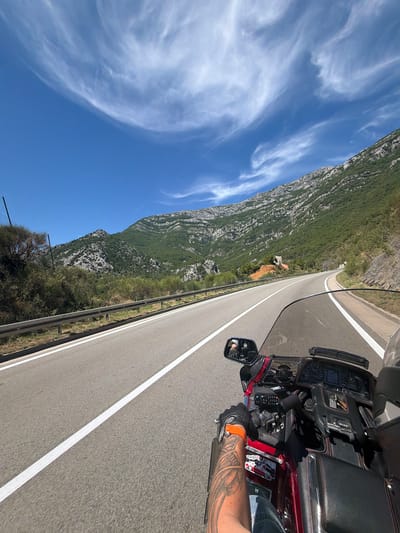 The image is a first-person perspective from a motorcycle traveling on a paved road in Đurmani, Montenegro. The road has white lane markings and a metal guardrail on the left. Steep, vegetated mountains with rocky outcrops rise on the right side of the road and in the background. The sky is bright blue with wispy white clouds. The motorcycle is a dark red touring model with a black leather seat and a large windshield. The rider's left arm, which is tattooed, is visible, and they are wearing a black glove with an orange accent. The motorcycle's dashboard with instruments is also visible. There are no other people clearly visible in the scene.