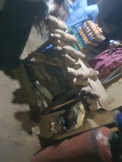 The image shows a street-side market scene in Dabawa, Nigeria. Several yams sit atop a rough wooden table or stand, likely for sale. Near the yams, there is a stack of egg cartons. Two people are partially visible; one wearing a light blue shirt, and another in a pink robe looking at a phone. Beside the stand is a bag of parboiled rice and a gas tank. The ground is wet.