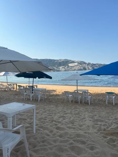 The image depicts a beachfront scene in Acapulco, Mexico. The view looks down on the beach, with white plastic chairs and tables arranged under various beach umbrellas. Some of the umbrellas feature the "Modelo" beer logo. In the background, across the water, there is a hillside covered in buildings. The sky is clear blue. The sand is light-colored and has numerous footprints and patterns. The water appears calm. There are no people visible in the shot.