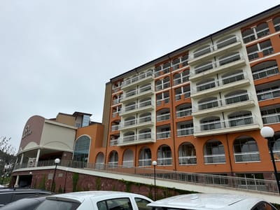 An eye-level, slightly low-angle exterior view captures a large, multi-story resort building under an overcast sky in Byala, Bulgaria. The building features a prominent orange facade on its right side, interspersed with light beige sections. This section is characterized by numerous white balconies with ornate, wavy railings, stacked in several rows, indicative of hotel rooms or apartments. Below these balconies, the lower levels of the orange facade feature large arched window openings, some also equipped with railings.

To the left, the building transitions to a more rounded, lighter-colored entrance structure with a large overhang supported by pillars. Above this entrance, the text "SOL PALM RESORT" is clearly visible, with "SOL" in larger, darker letters. A long, gently sloping ramp with silver metal handrails runs along the front of the property, ascending towards the main entrance. The retaining wall bordering the ramp is made of reddish-brown stone, partially covered with climbing ivy. Several black lampposts with spherical white lights are spaced along the ramp.

In the immediate foreground, the tops and hoods of several parked cars, including white, black, and grey vehicles, are visible. The surfaces of these cars appear to have water droplets, suggesting recent rain or mist. The sky is a uniform light grey, indicating a cloudy or overcast day, and there are no visible people or specific activities taking place, creating a quiet, still atmosphere.