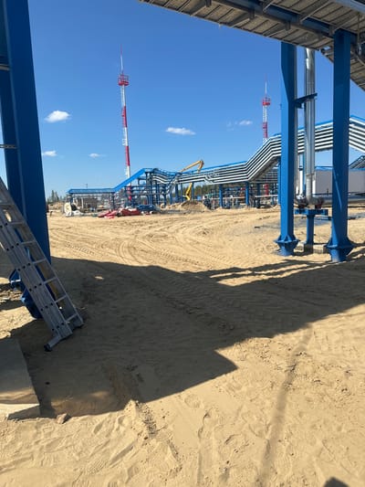 The image depicts an outdoor industrial construction site under a clear blue sky with scattered clouds. The foreground is dominated by sandy ground with tire tracks and some scattered debris. A silver ladder leans against a large blue metal support structure on the left side of the frame.
In the mid-ground, an extensive network of blue metal structures and elevated pipelines is visible. Two tall, red and white striped towers with observation platforms and antennas stand in the background. A yellow construction excavator is partially visible among the structures. The ground throughout the site appears to be sand and dirt, indicating ongoing construction or site preparation. The overall impression is one of a large-scale industrial project in progress. There are no people visible in the image.