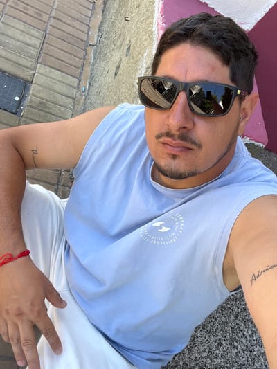 A single male individual is depicted in a high-angle, close-up outdoor photograph taken in Bernal Este, Argentina. The subject has dark, short hair, a mustache, and a short beard. He wears dark, reflective rectangular sunglasses, a light blue sleeveless shirt with a white circular logo containing text and a wave graphic, and light-colored pants. A red string bracelet is present on his left wrist. Tattoos include the word "Romantic" in script on his right bicep and "Adria" in script on his left bicep. A silver stud earring is visible in his left earlobe. The background features a brick-paved ground and a wall with visible patches of pink and dark red paint. Reflections in the sunglasses show building structures and foliage. The scene is illuminated by direct daylight.