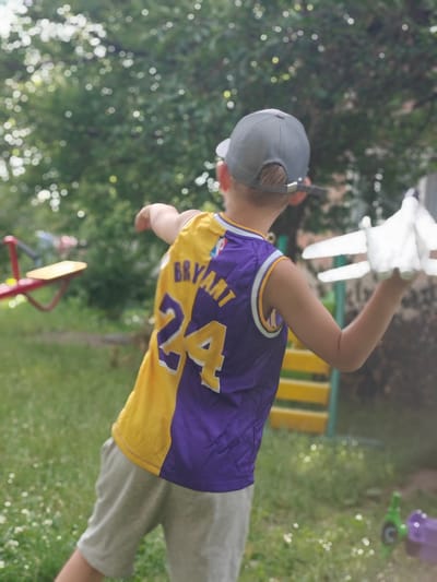 A young boy, viewed from behind, is standing in a grassy outdoor area. He is wearing a yellow and purple basketball jersey with the name "BRYANT" and the number "24" visible on the back. He also has on a gray baseball cap and light gray shorts. In his right hand, he is holding a white toy airplane.
The background suggests a playground setting, with glimpses of a red seesaw on the left and a yellow structure with green supports to the right. The overall scene is outdoors with green trees and grass, indicating a daytime setting with ample natural light, likely sunny given the bright highlights. The soft focus of the background suggests the image was taken with a shallow depth of field, drawing attention to the boy and his toy. The mood appears playful and relaxed. The location is identified as Hadiach, Ukraine.