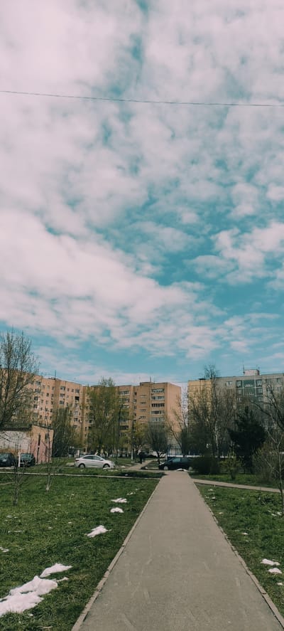 A tilted, outdoor shot shows a street scene in Fryazino, Russia. The sky occupies the left half of the image, displaying a bright blue hue dotted with white clouds. A power line is visible. On the right side, apartment buildings with many windows. The street's sidewalk is grey and there is a patch of green grass. There are small spots of melting snow. Cars are parked along the street and bare trees are visible. The perspective creates a sense of depth leading into the image.