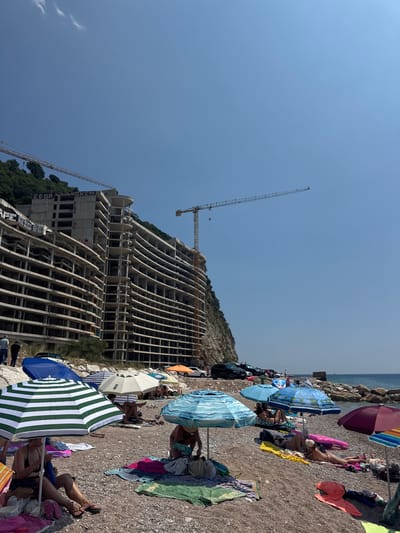 In Mirište, Montenegro, a beach scene unfolds under a clear blue sky. Construction cranes loom over a partially built, multi-story concrete structure, which appears to be abandoned and covered in graffiti. Below, on a pebbly beach, beachgoers relax under various umbrellas. Several people are seated or lying on towels, some appearing to sunbathe. The sea is visible in the background, with a rocky shoreline. Cars are parked along the edge of the beach, adjacent to the construction site.