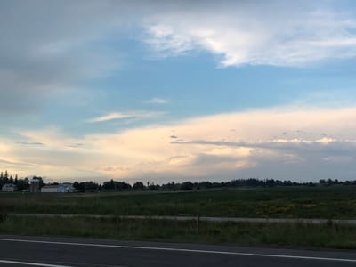 This media file captures a pastoral scene in Bradford West Gwillimbury, Canada, likely during the late afternoon or early evening given the soft, diffused lighting. The sky is a canvas of dynamic clouds, transitioning from a pale blue to warm, peachy hues where sunlight filters through. Wispy white clouds drift across the upper portion of the frame, while larger, more textured formations dominate the mid-ground, suggesting approaching or receding weather.
The landscape below is a patchwork of agricultural fields. In the distance, a farmstead is visible, featuring a prominent silo and several buildings, including a long, low-rise structure that may be a barn or workshop. The fields are a mix of green crops, with some areas appearing darker and more lush, and others lighter, possibly indicating different stages of growth or crop types. A line of trees, including evergreens, borders the fields and creates a sense of depth.
In the foreground, a paved surface, likely a road, is partially visible with white lane markings. A fence runs along the edge of the field, separating it from the roadside. The overall impression is one of tranquility and rural life, with a gentle, somewhat dramatic sky setting a contemplative mood. There is no visible text in the image.