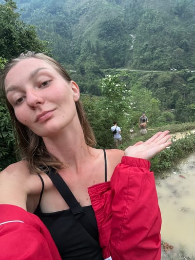 A young woman is featured prominently in the foreground of this vertical outdoor shot, with a lush, mountainous landscape stretching behind her. She is positioned on the left side of the frame, looking towards the viewer with a neutral expression. She wears a black top with thin straps, partially covered by a vibrant red jacket that drapes over her left arm and torso. Her right hand is raised, palm slightly open, in front of her face, with the forearm of her red jacket visible.
The background reveals dense, verdant foliage covering steep slopes and hills, suggesting a forest or jungle environment typical of Nậm Dịch Commune, Vietnam. A narrow, unpaved path or dirt track winds through the mid-ground, where two other individuals are visible. One person, appearing to be a man, is seen carrying a small, light-colored animal, possibly a dog or goat, while another person walks slightly behind him. Further into the background, a body of murky, light-brown water is visible, bordered by more greenery, which could be a flooded rice paddy or a natural pond. The lighting suggests it is daytime, possibly under an overcast sky, given the soft, diffused light and lack of harsh shadows. No text is visible in the image. The scene captures an outdoor activity, likely hiking or trekking, amidst the natural beauty of the Vietnamese mountains.