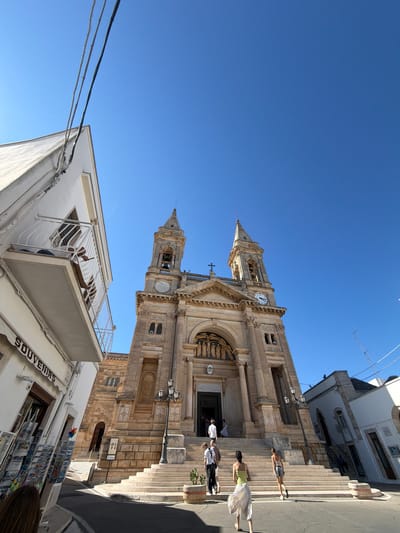 The image shows a view of the Basilica Santuario della Madonna della Madia in Monopoli, Italy. The basilica is a large, stone church with two bell towers and a clock. It is located in a city square with a few people walking around. The sky is blue and clear. The Basilica is a popular tourist destination and is known for its stunning architecture. The church is a beautiful example of Baroque architecture, and its facade is decorated with intricate sculptures and carvings. The interior of the basilica is also impressive, with a large nave, a dome, and a number of chapels. The church is an important religious site and is visited by many pilgrims each year.