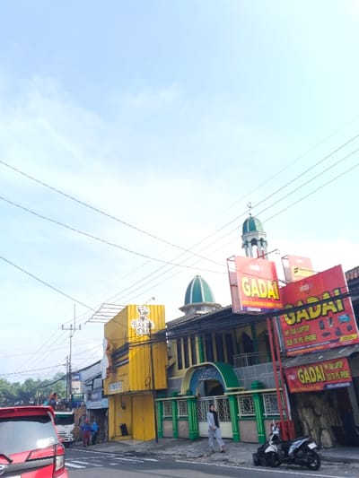 A street scene depicts a yellow building with "BAKER OLD" painted on its side next to a mosque adorned with green accents and domes. A woman in a hijab walks in front of the mosque. Signs advertising "GADAI" (pawn shop) with phone repair services for various devices are displayed. A red car is visible on the left side of the frame, and a motorcycle is parked near the right. Power lines crisscross against a pale sky in the background.