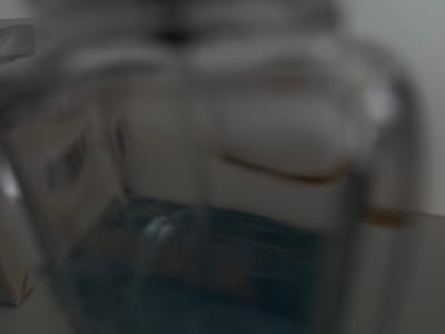The image shows a close-up, out-of-focus view of a clear glass bottle containing a blue liquid. The bottle is partially filled, with the liquid occupying the lower portion. The background is blurred and appears to be a light-colored surface. There is a faint barcode-like pattern visible on a label attached to the bottle. The overall impression is one of simple object photography, with no discernible activity or specific setting beyond the bottle itself. The lighting is soft, and there are no indicators of time of day or weather conditions. The image is devoid of any human presence or emotional context. The location context provided indicates Mülheim an der Ruhr, Germany, but this information is not visually verifiable from the image itself.