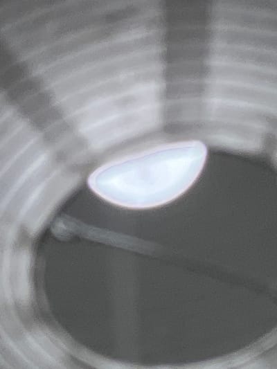 The image displays a close-up, abstract view of a reflective, iridescent object against a muted, striped background. The object, appearing oval-shaped and luminescent with soft purples and blues, is centrally located and slightly out of focus. The background consists of concentric, blurred grey and white lines, suggesting a ribbed or layered surface. No specific location in Буран Снт, Russia, can be identified from this image. The scene is still and devoid of any human presence or activity.